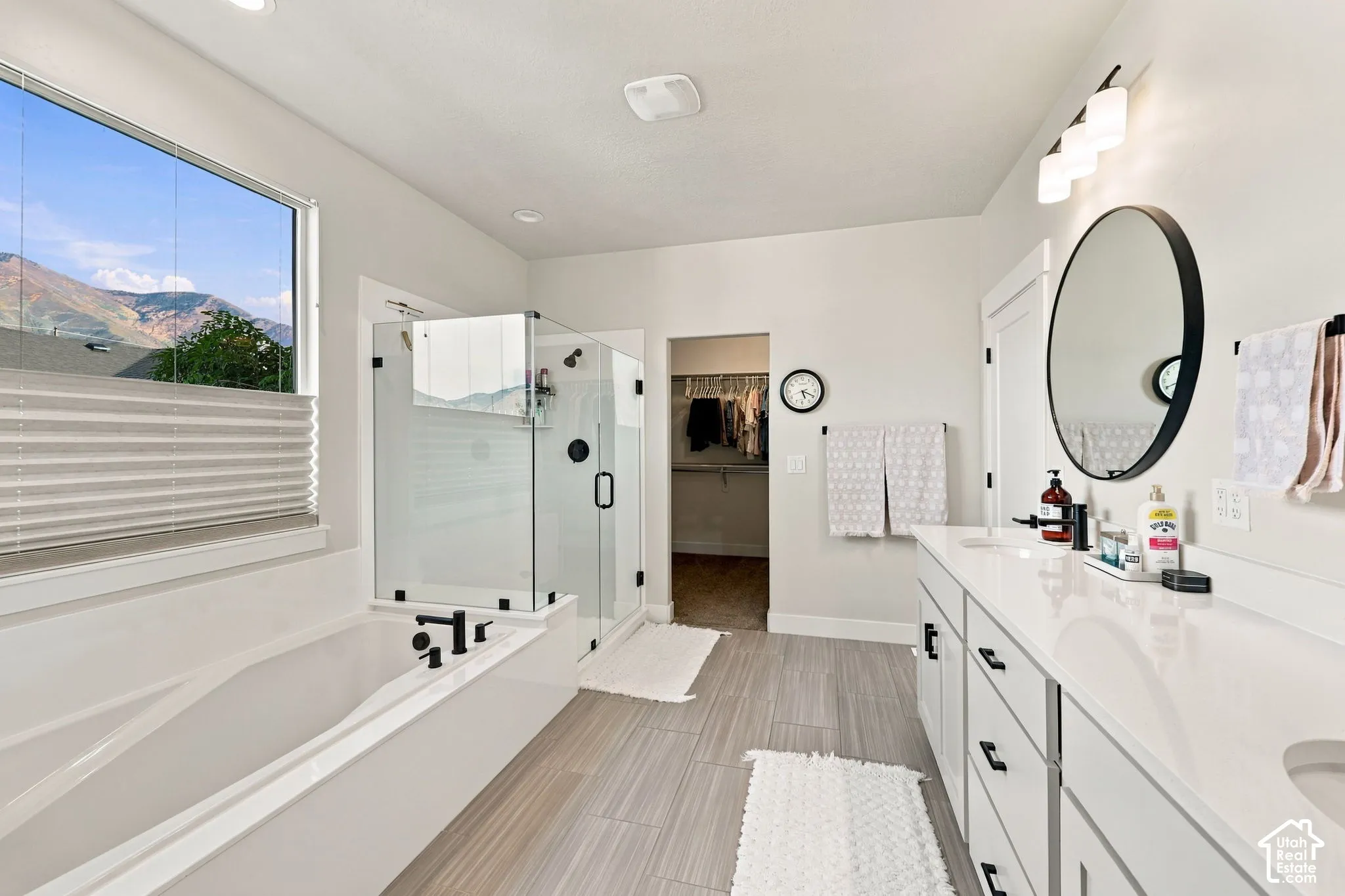 Soaking tub with black fixtures, walk-in glass shower, dual vanity with white countertop, round mirror, and wood-finish flooring