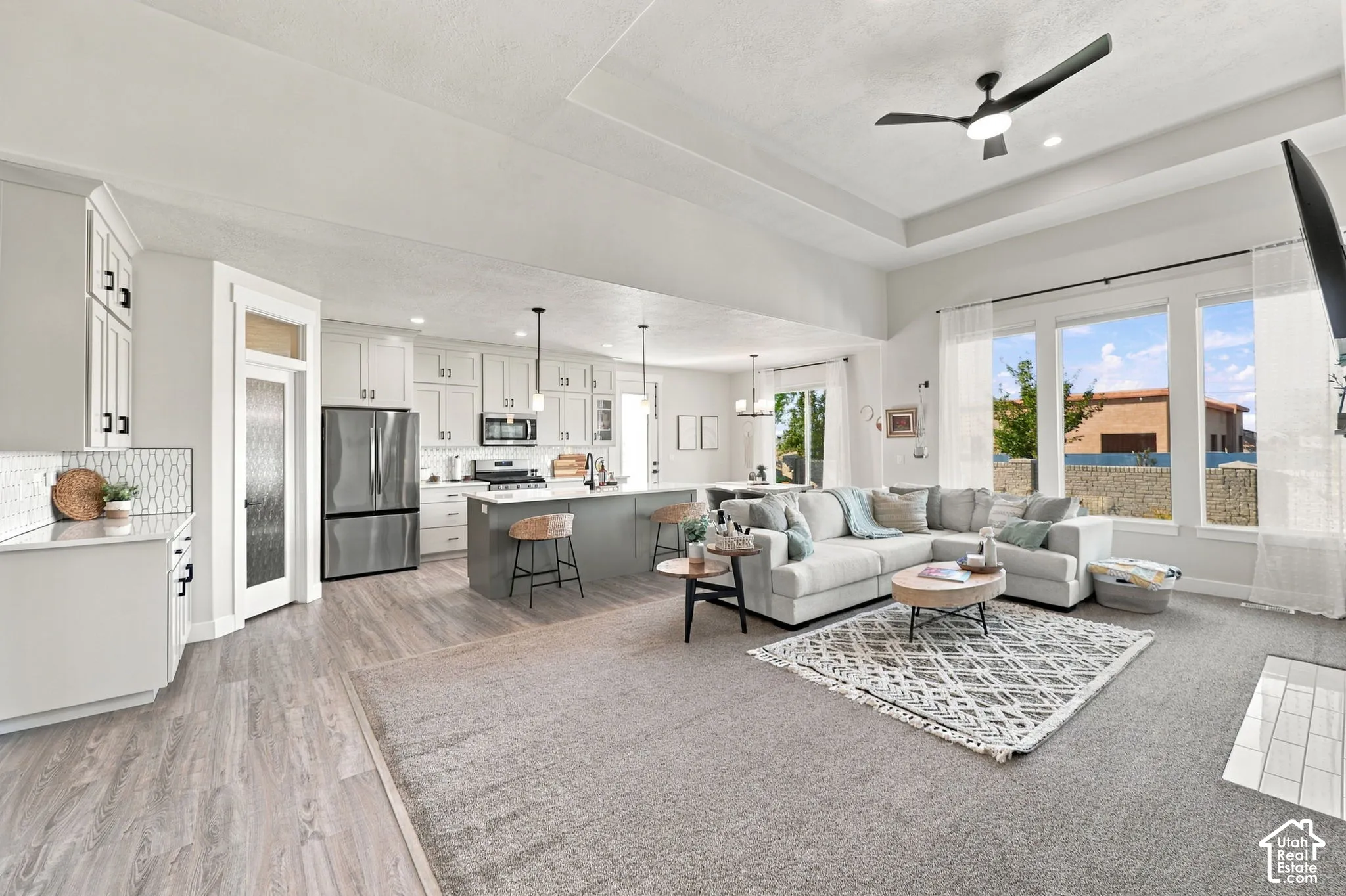 Open-concept living space featuring a spacious kitchen, built-in stainless steel appliances, large windows, and a tray ceiling with a modern ceiling fan