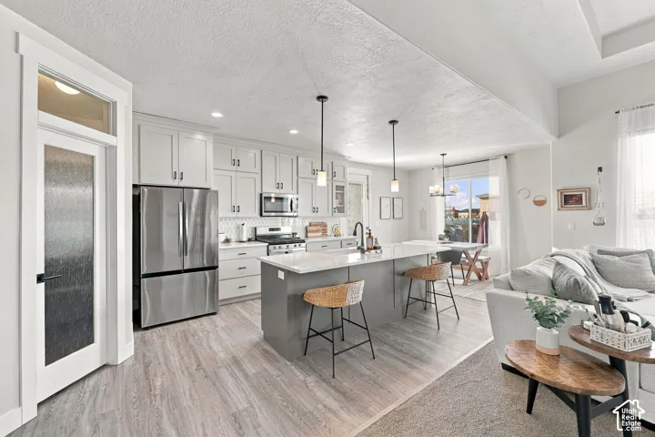 Open-concept kitchen featuring light-toned cabinetry, stainless steel appliances, a center island with pendant lighting, and wood-finish flooring