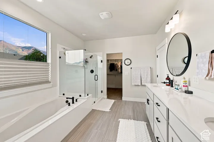 Soaking tub with black fixtures, walk-in glass shower, dual vanity with white countertop, round mirror, and wood-finish flooring