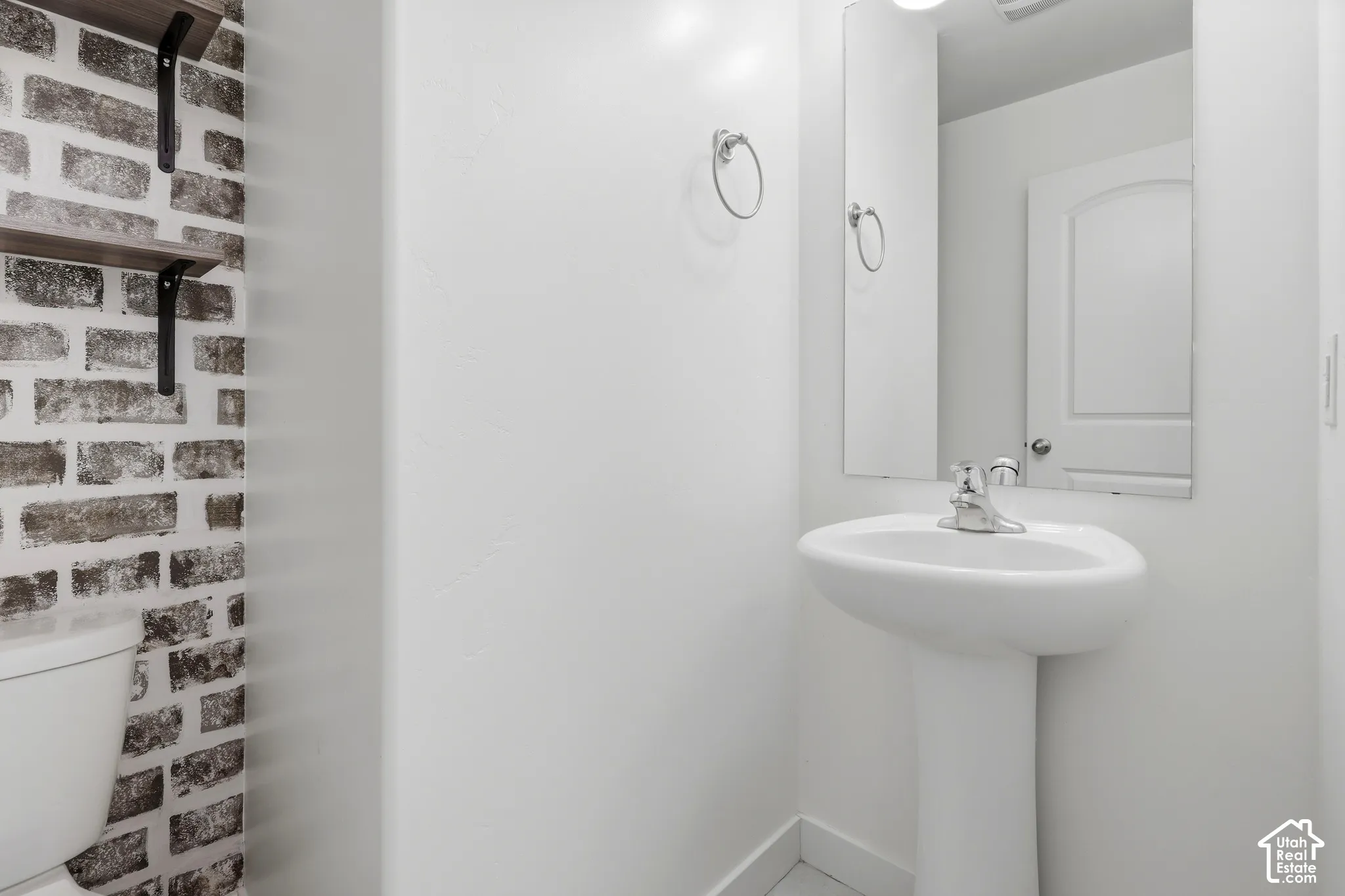 Pedestal sink with chrome faucet, wall-mounted mirror, and decorative brick-patterned wall
