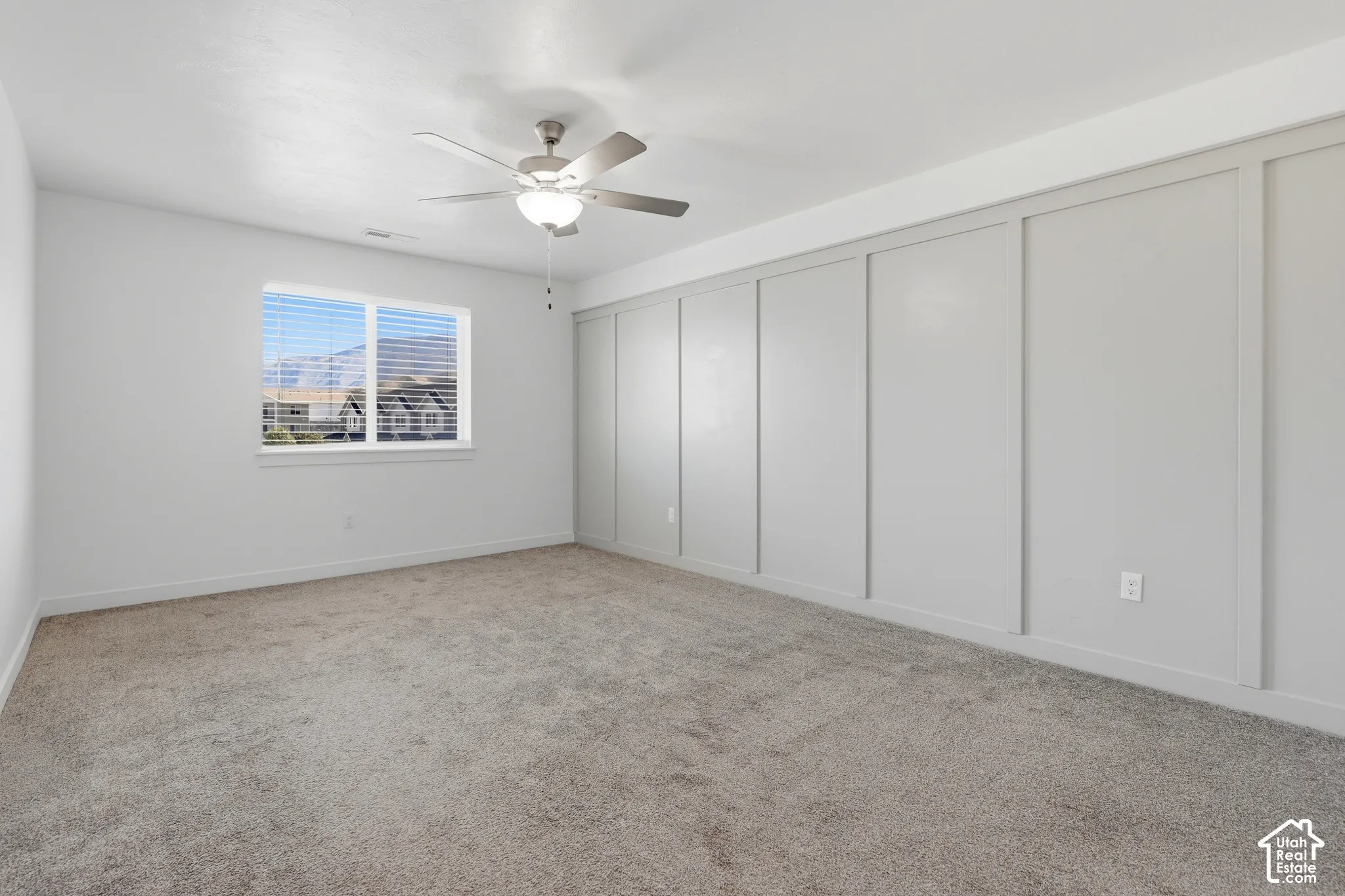 Neutral-toned carpeting, ample natural light from a large window, a ceiling fan with integrated lighting, and a feature wall with decorative paneling