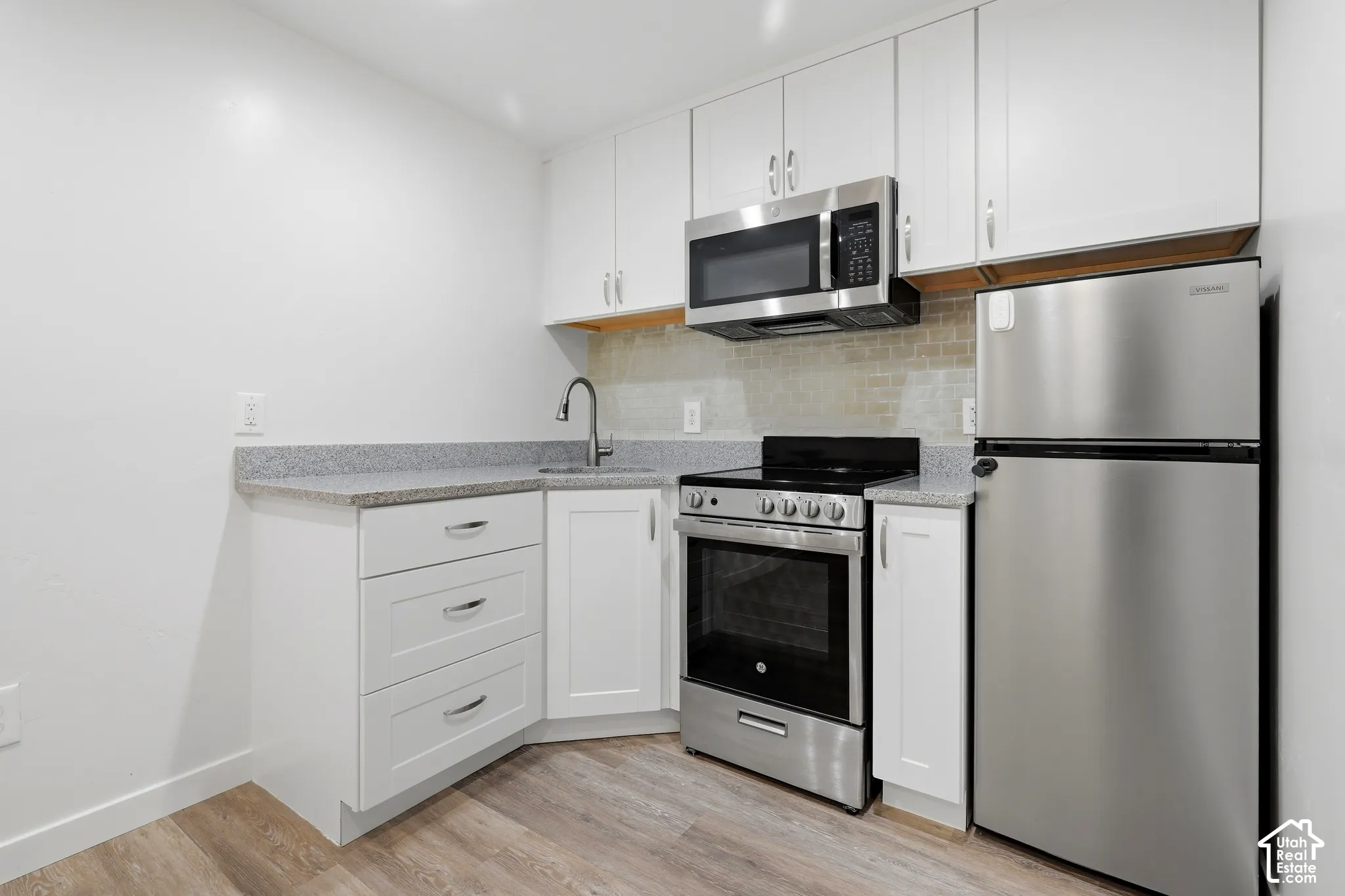 White cabinetry, light-toned wood-finish flooring, stainless steel appliances, granite-style countertops, and a tile backsplash