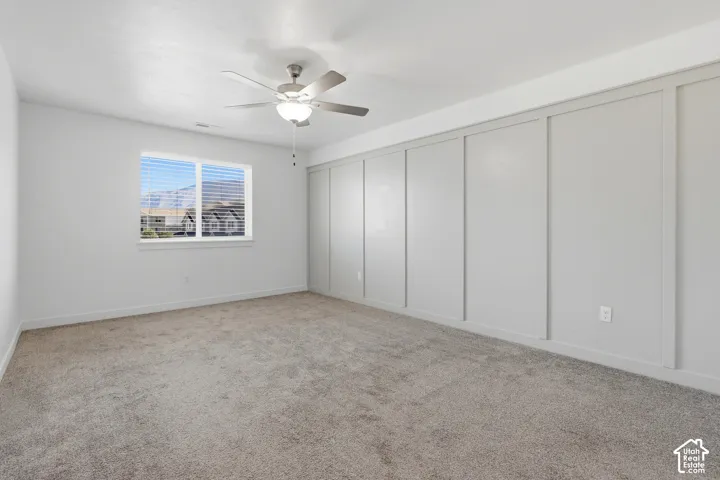 Neutral-toned carpeting, ample natural light from a large window, a ceiling fan with integrated lighting, and a feature wall with decorative paneling