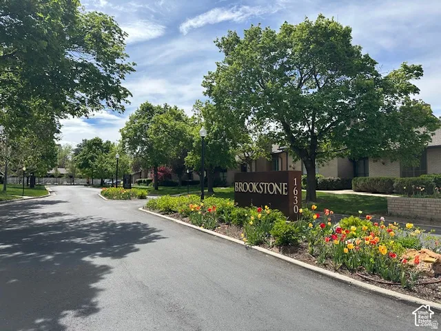 Asphalt paved road with mature trees and a "BROOKSTONE 1630" sign, featuring landscaped flower beds with colorful blooms