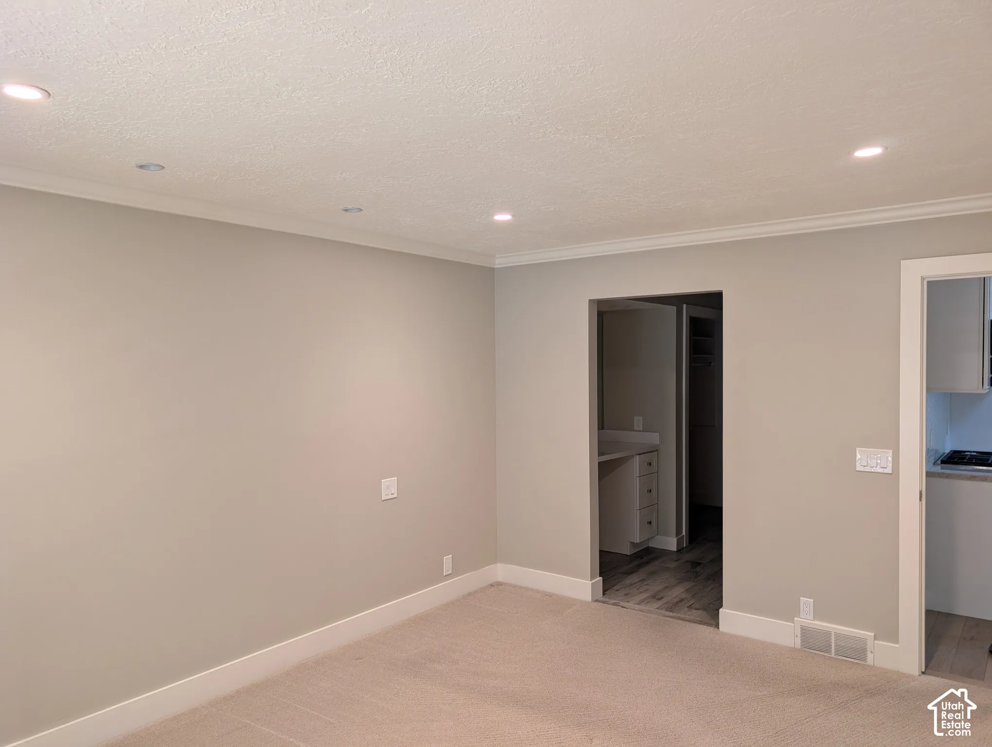 Recessed lighting, crown molding, light-toned wall paint, carpeted flooring, and a doorway to an adjoining area with wood-finish flooring
