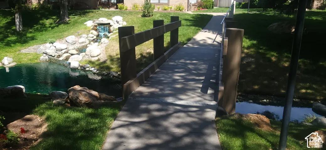 Concrete pedestrian bridge with wood-finish railings, surrounded by a manicured lawn, a rock-lined pond, and a multi-tiered rock waterfall feature