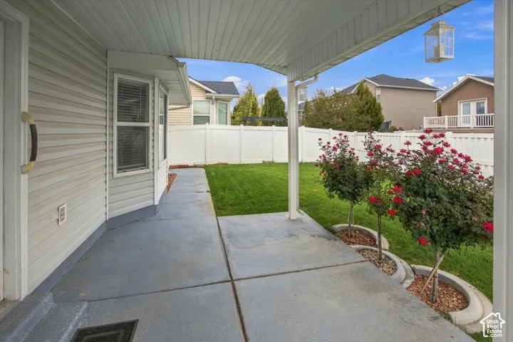 Covered patio with a concrete slab, white vinyl siding, bay window, landscaped planting beds with flowering shrubs, and a fenced backyard