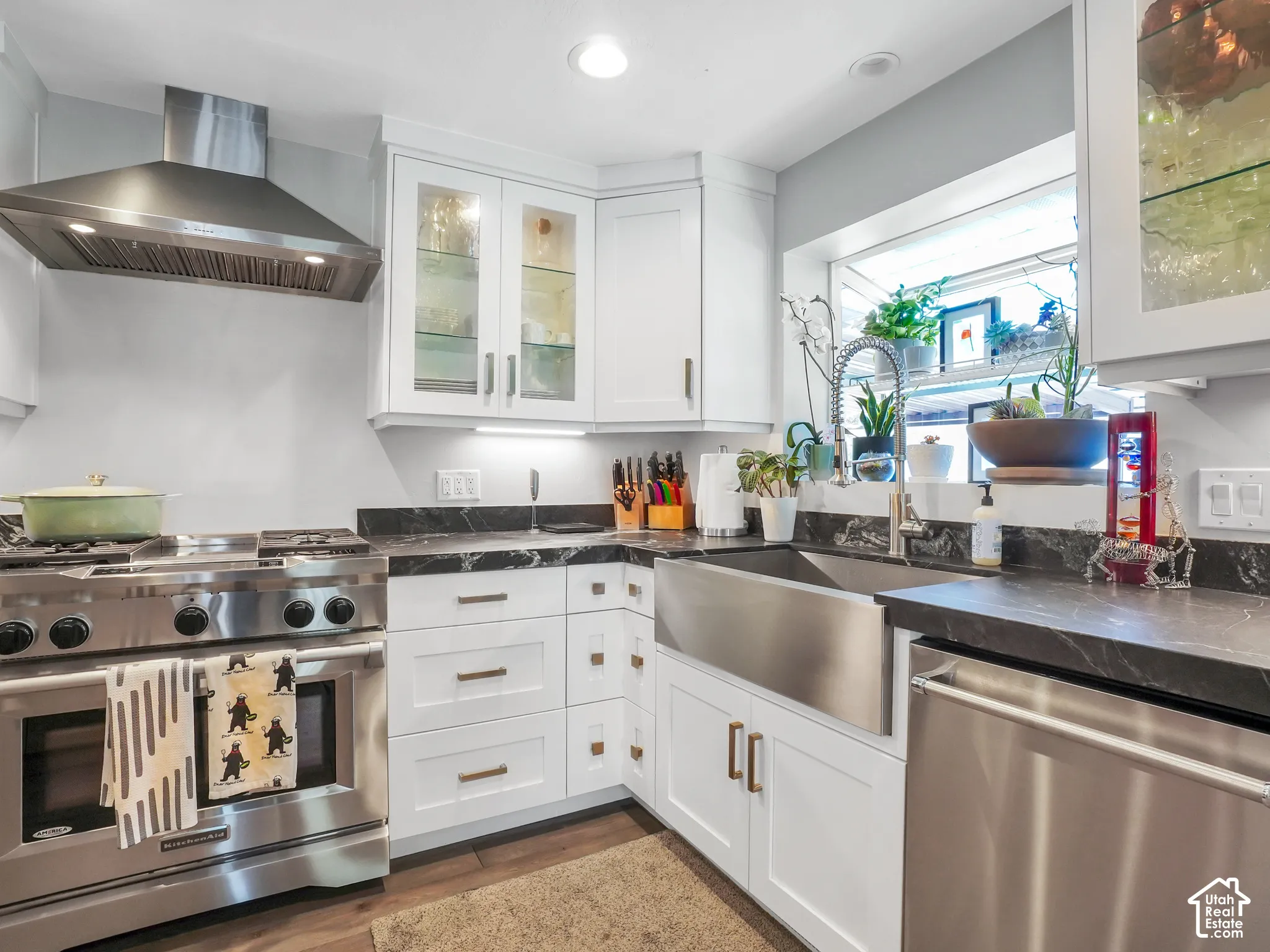 White cabinetry with glass-front uppers, stainless steel range with hood, dark countertops, stainless steel farmhouse sink, and stainless steel dishwasher
