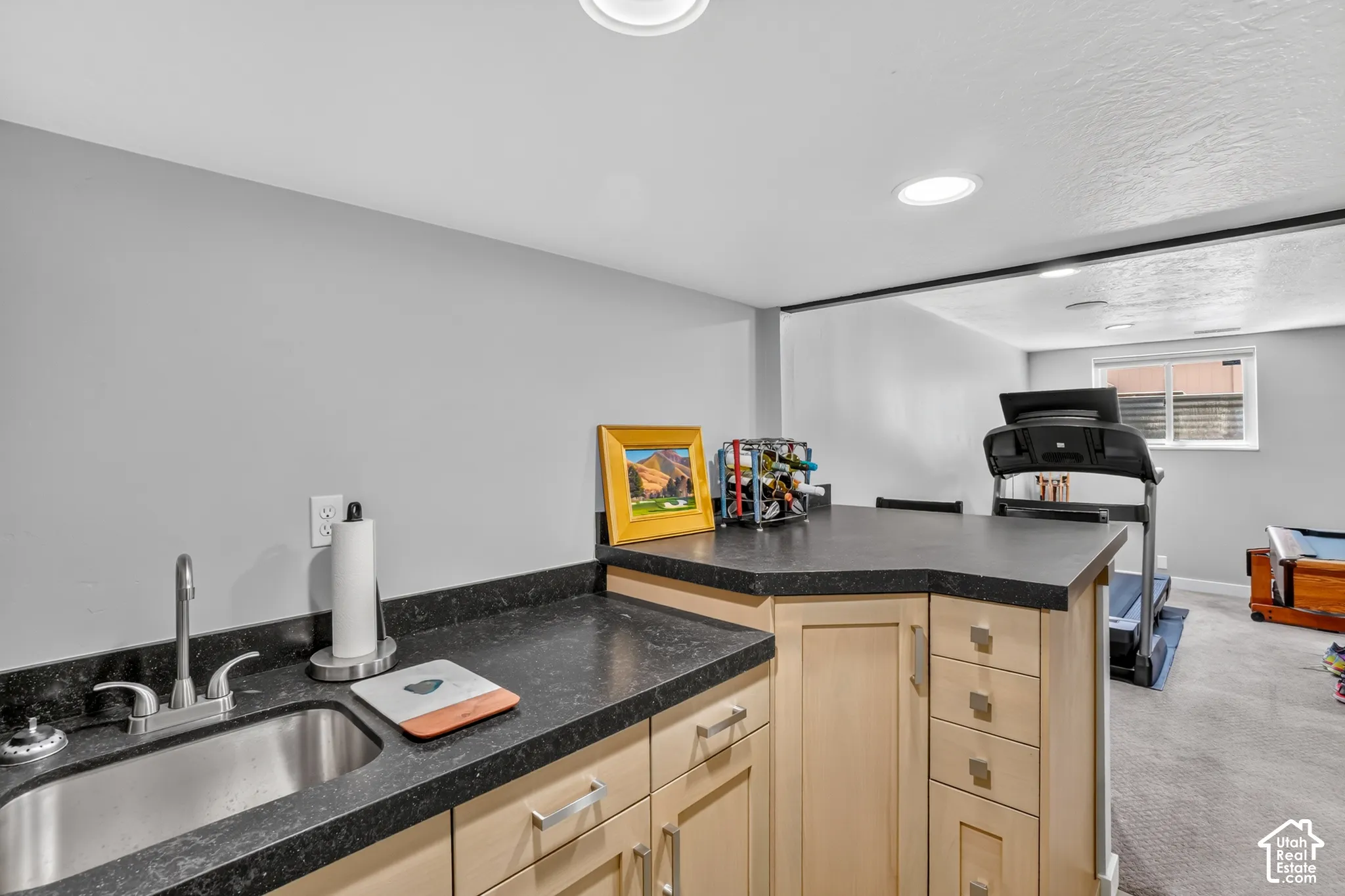 Integrated wet bar with stainless steel sink, dark-toned countertop, and light wood-finish cabinetry