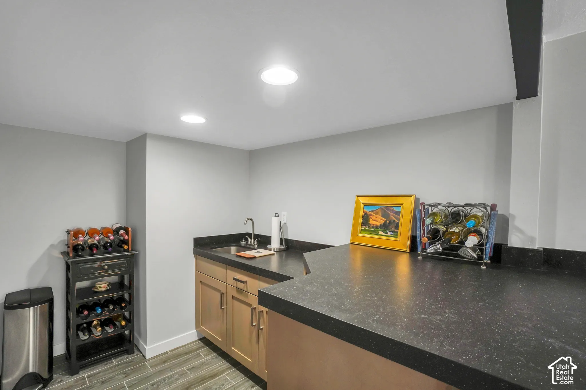 Wet bar featuring a stainless steel sink, wood-finish cabinetry, dark countertops, and recessed lighting