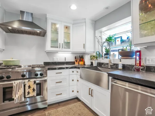 White cabinetry with glass-front uppers, stainless steel range with hood, dark countertops, stainless steel farmhouse sink, and stainless steel dishwasher