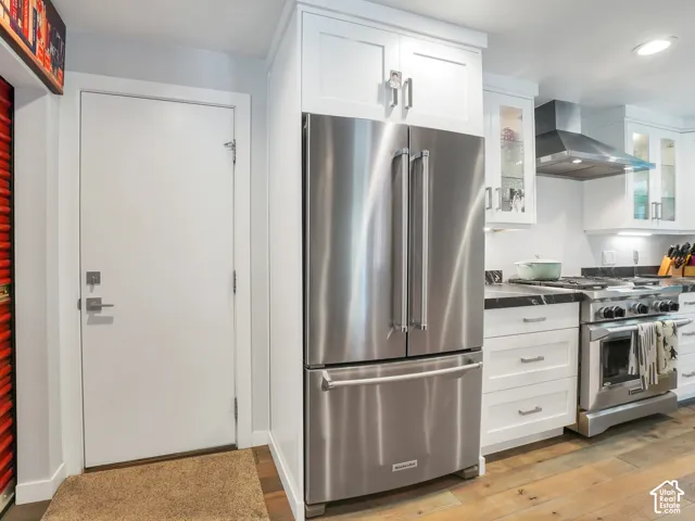Stainless steel French door refrigerator, white shaker-style cabinetry with glass accents, stainless steel range with hood, dark-toned countertops, and wood-finish flooring