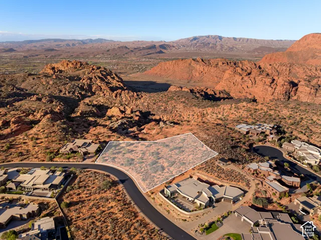 Expansive desert lot featuring a curved paved road, surrounding residential structures, and natural rock formations