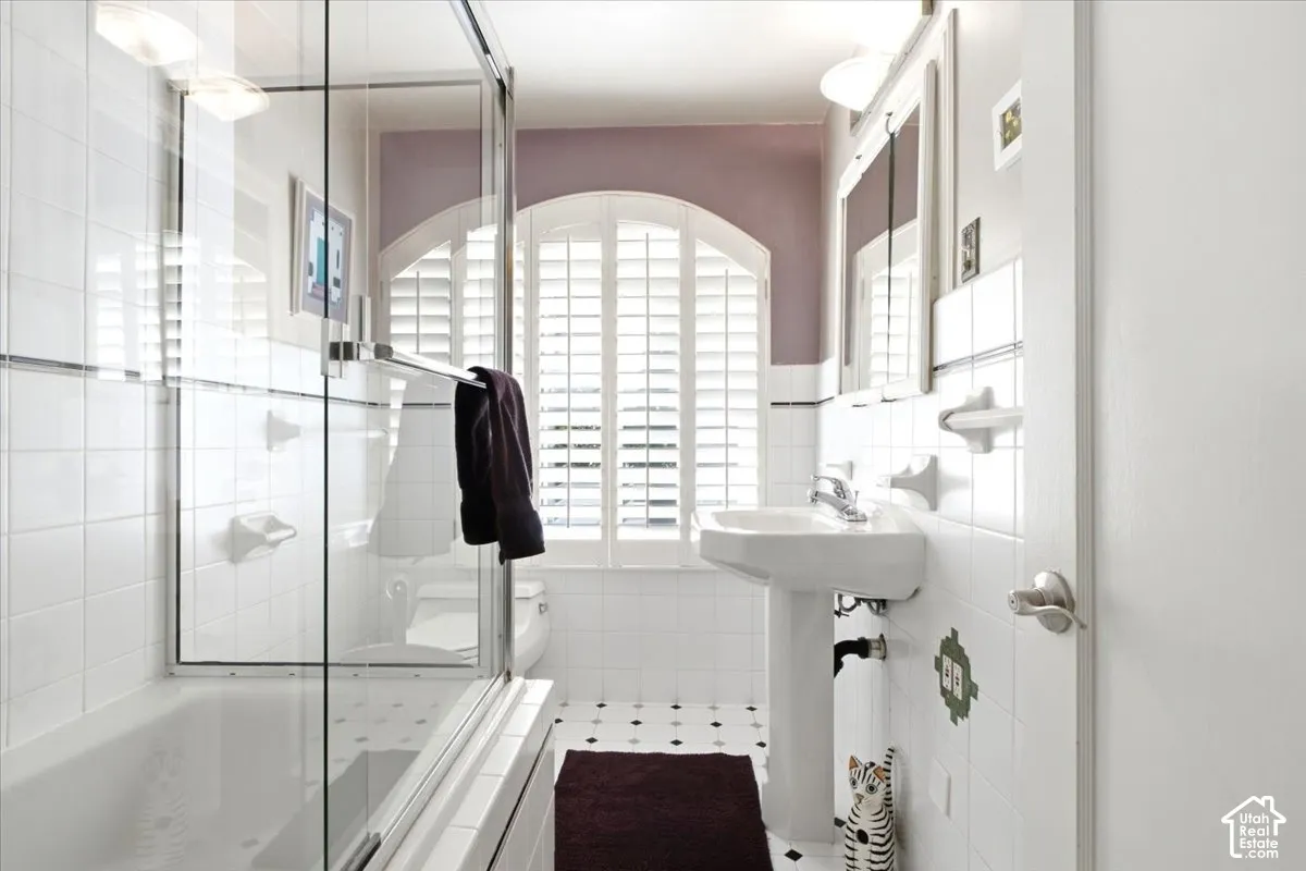Tiled bathroom featuring a glass-enclosed shower/tub combination, pedestal sink, and arched window with plantation shutters