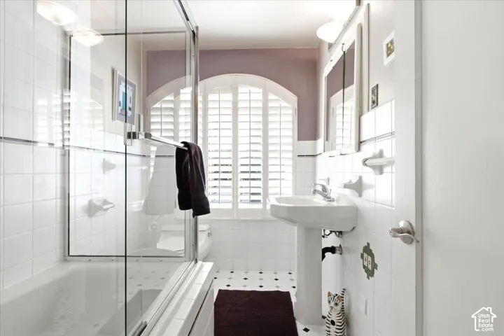 Tiled bathroom featuring a glass-enclosed shower/tub combination, pedestal sink, and arched window with plantation shutters