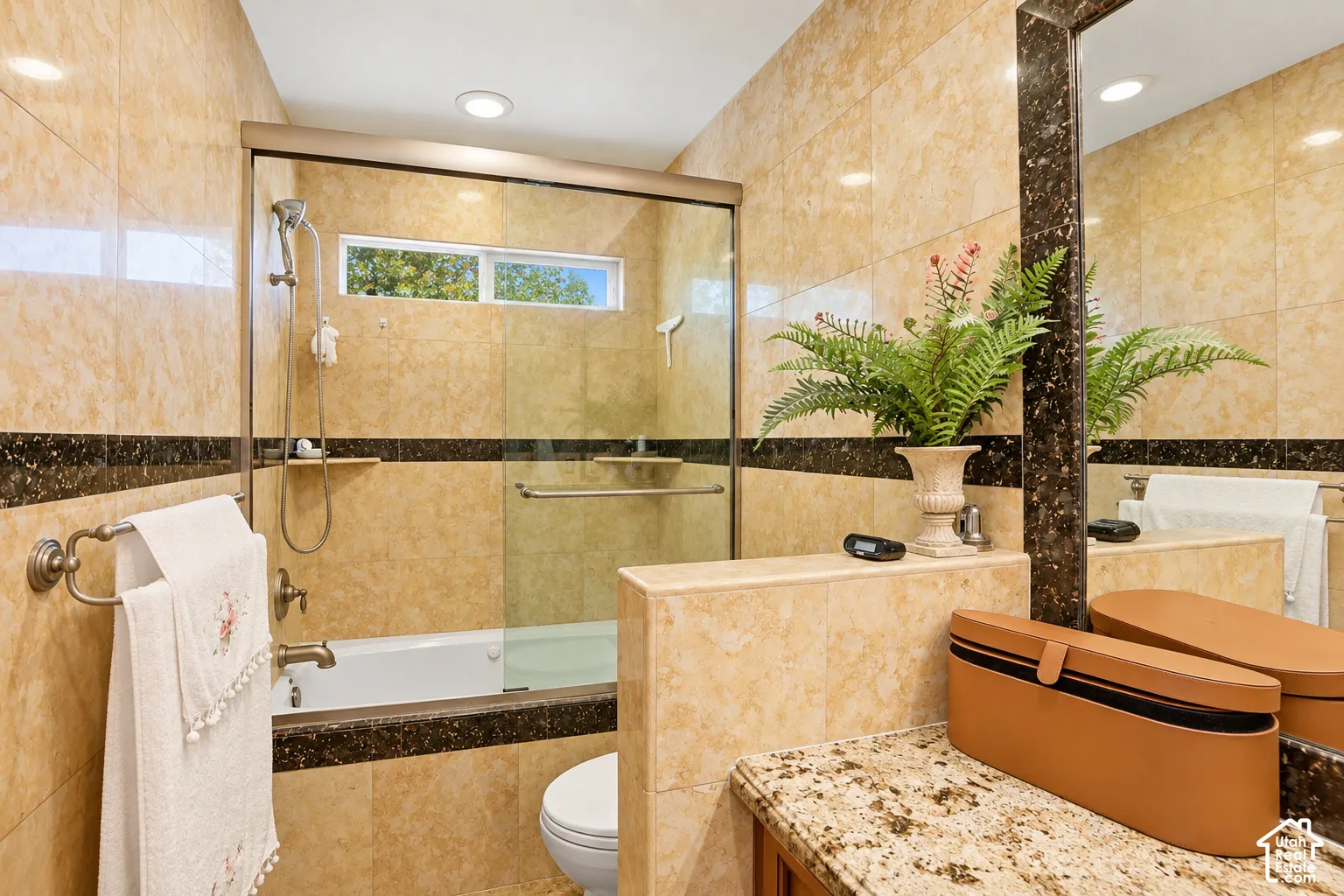 Full-height tiled walls, glass-enclosed shower over a built-in tub, transom window, recessed lighting, and a granite-finish vanity