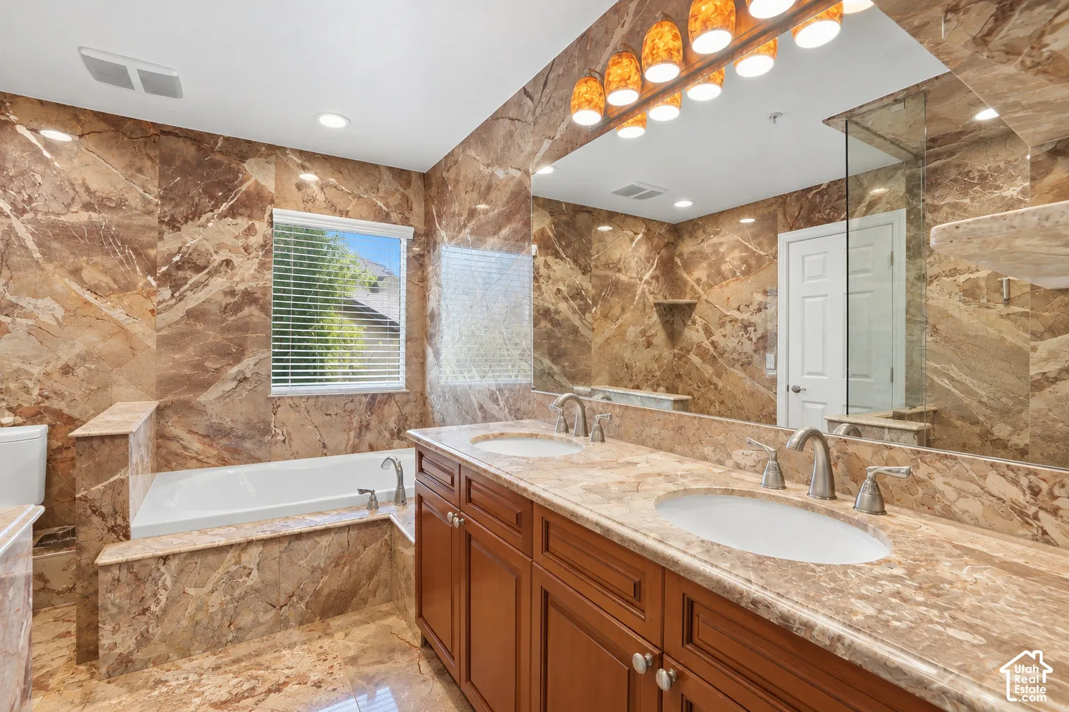 Bathroom featuring extensive natural stone tiling, a built-in soaking tub, a dual vanity with undermount sinks, and a large wall-mounted mirror