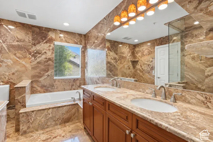 Primary en suite bathroom featuring extensive stone tiling, a built-in soaking tub, a dual vanity with undermount sinks, and a large wall-mounted mirror