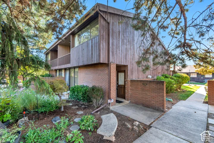 Brick and wood-paneled exterior with a front entry, featuring an upper-level balcony, ground-level windows, and a landscaped garden area