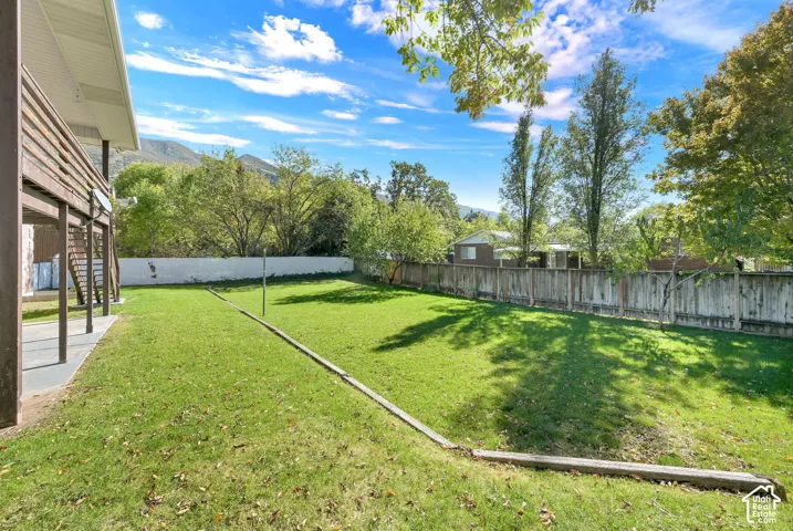 Expansive backyard featuring a manicured lawn, perimeter wood fencing, tiered landscape edging, a covered concrete patio, and mature trees