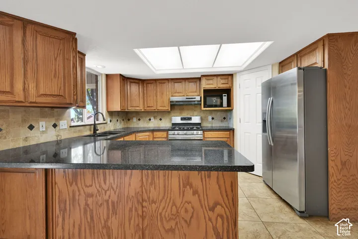 Kitchen featuring wood cabinetry, dark stone countertops, natural stone tile backsplash, stainless steel appliances, and a large skylight