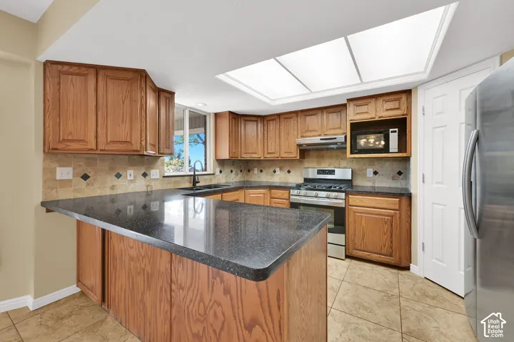 Kitchen featuring wood cabinetry, dark solid-surface countertops, tile backsplash with inset accent tiles, a skylight, and stainless steel appliances