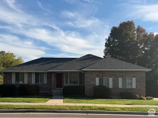 Brick-clad exterior with a dark roof, front yard with manicured lawn, concrete walkway, and mature trees