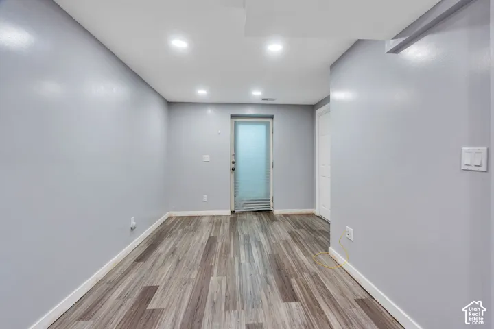 Spacious room featuring wood-finish flooring, recessed lighting, neutral painted walls, and a white interior door