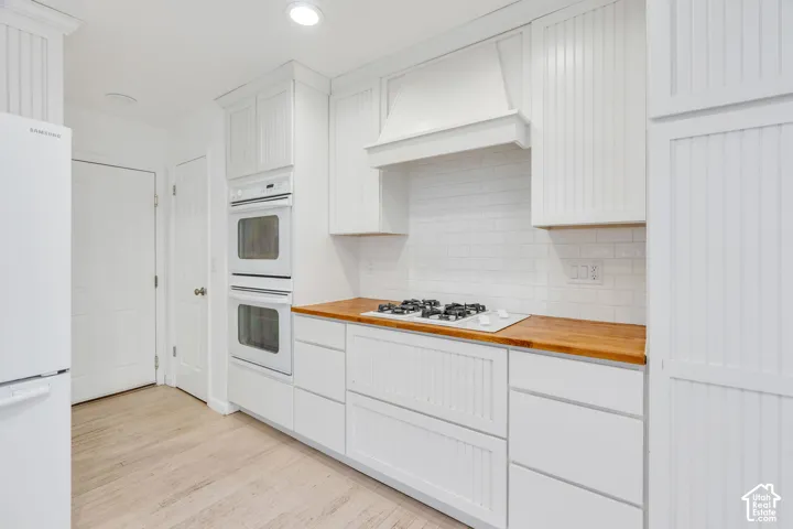White beadboard cabinetry, double wall ovens, wood-finish countertops, white subway tile backsplash, and light wood-finish flooring