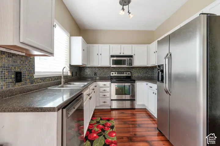 White cabinetry with dark hardware, stainless steel appliances, dark countertops, mosaic tile backsplash, and wood-finish flooring