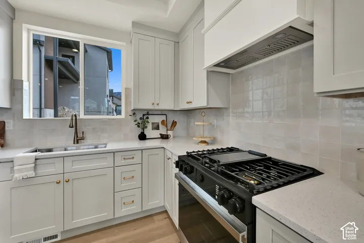 White cabinetry with gold-tone hardware, white stone countertops, stainless steel sink with bronze-finish faucet, white subway tile backsplash, and a black gas range