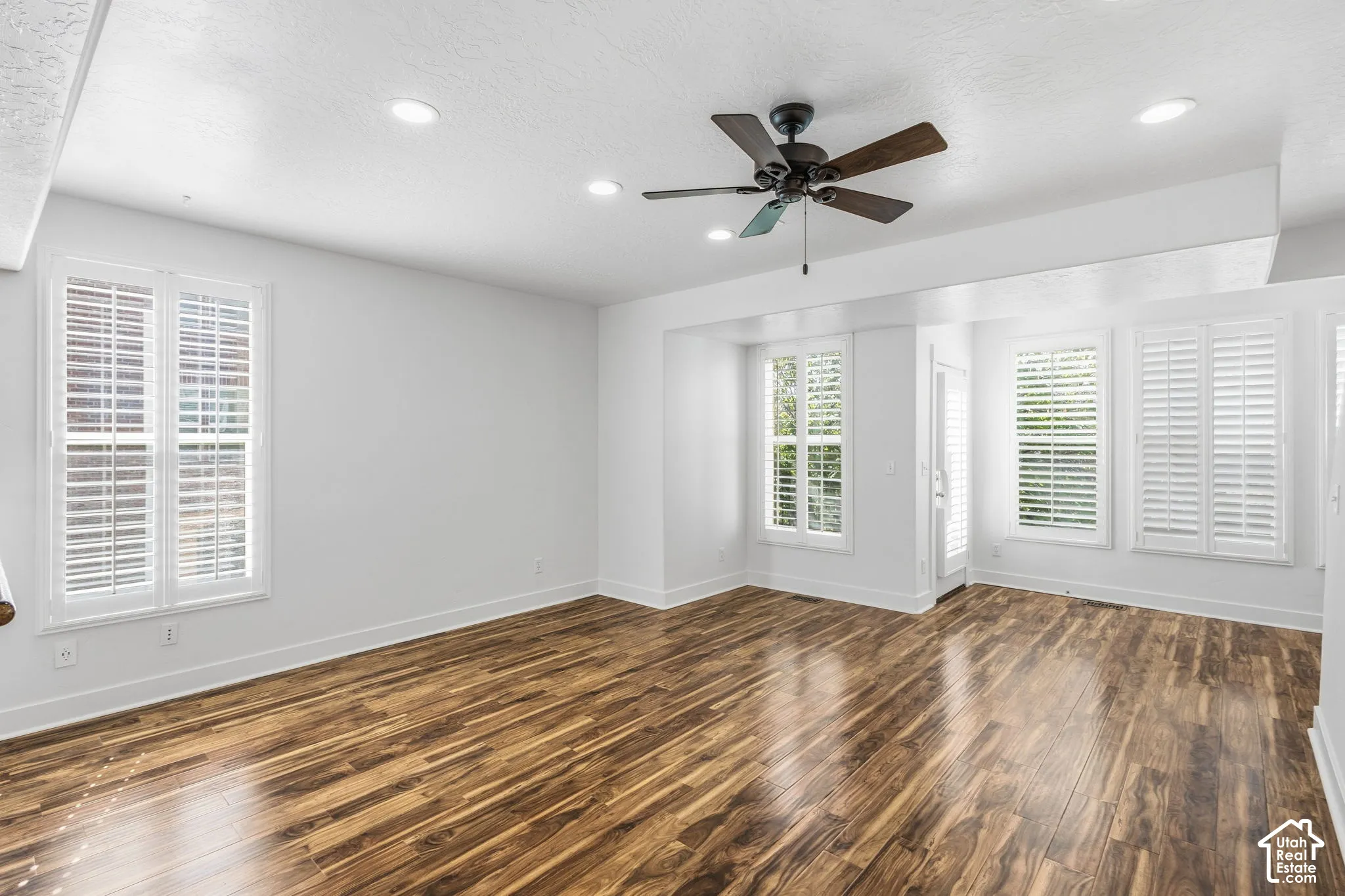 Spacious room featuring wood-finish flooring, white walls, multiple windows with white plantation shutters, a ceiling fan, and recessed lighting