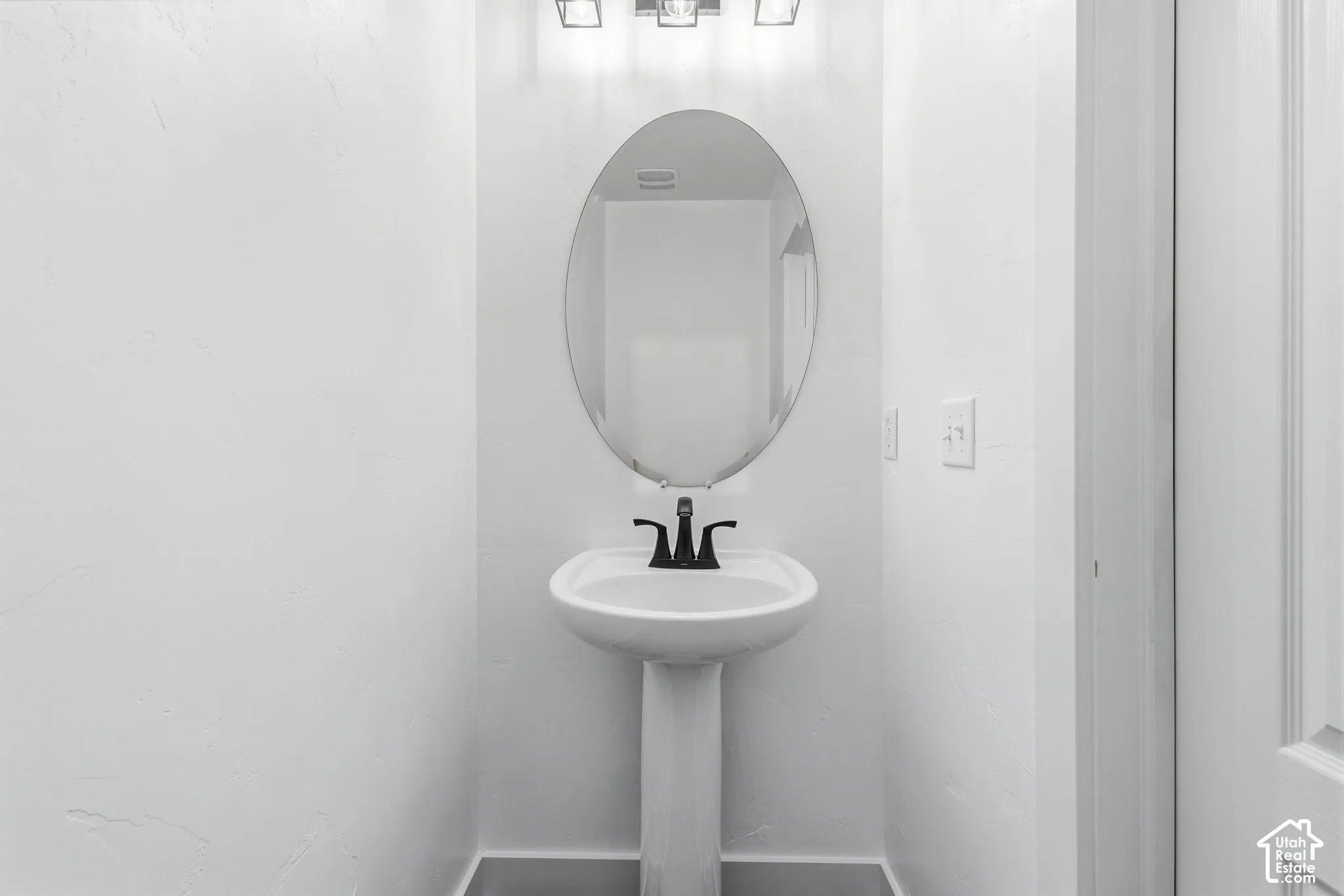 Pedestal sink with a black faucet, oval mirror, and a three-light vanity fixture