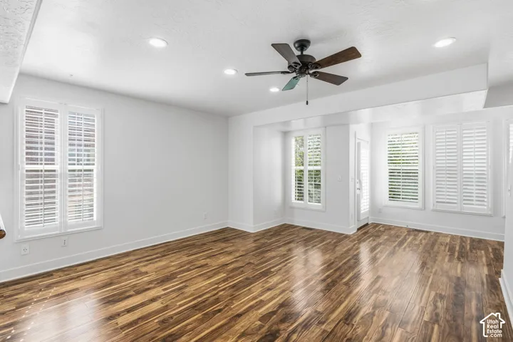 Spacious room featuring wood-finish flooring, white walls, multiple windows with white plantation shutters, a ceiling fan, and recessed lighting