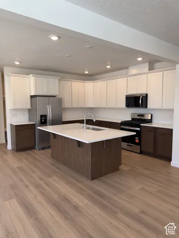 Kitchen featuring white upper cabinetry, wood-finish lower cabinetry, a center island with sink, stainless steel appliances, and wood-finish flooring
