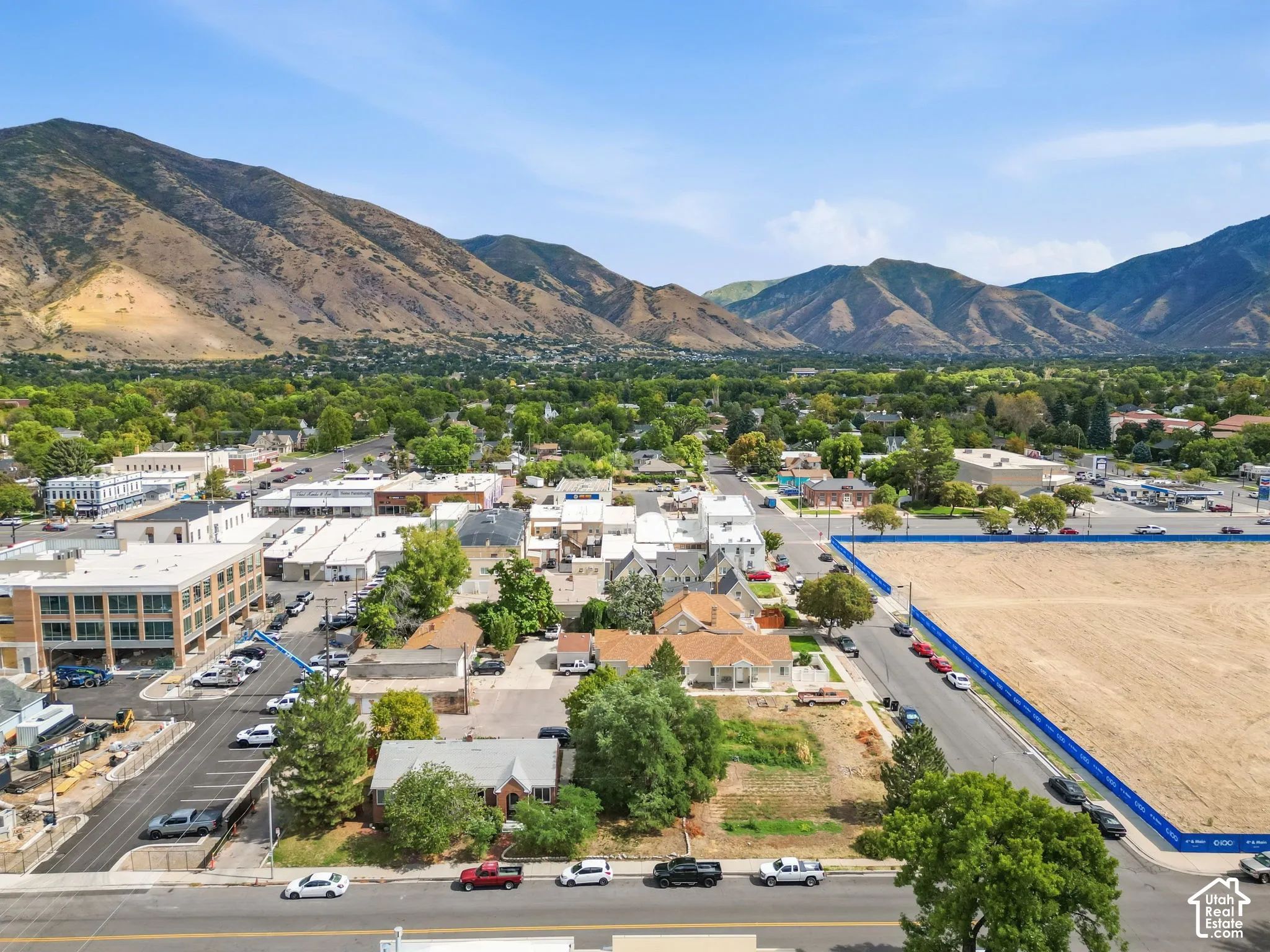 Aerial view showcasing this property's prominent corner lot positioning with direct street frontage, mature landscaping throughout the surrounding grounds, and proximity to the riverfront and adjacent commercial district