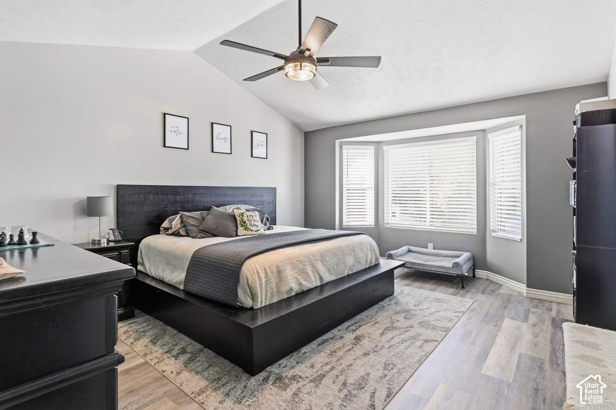Vaulted ceiling with a ceiling fan, bay window with blinds, wood-finish flooring, and black bedroom furniture