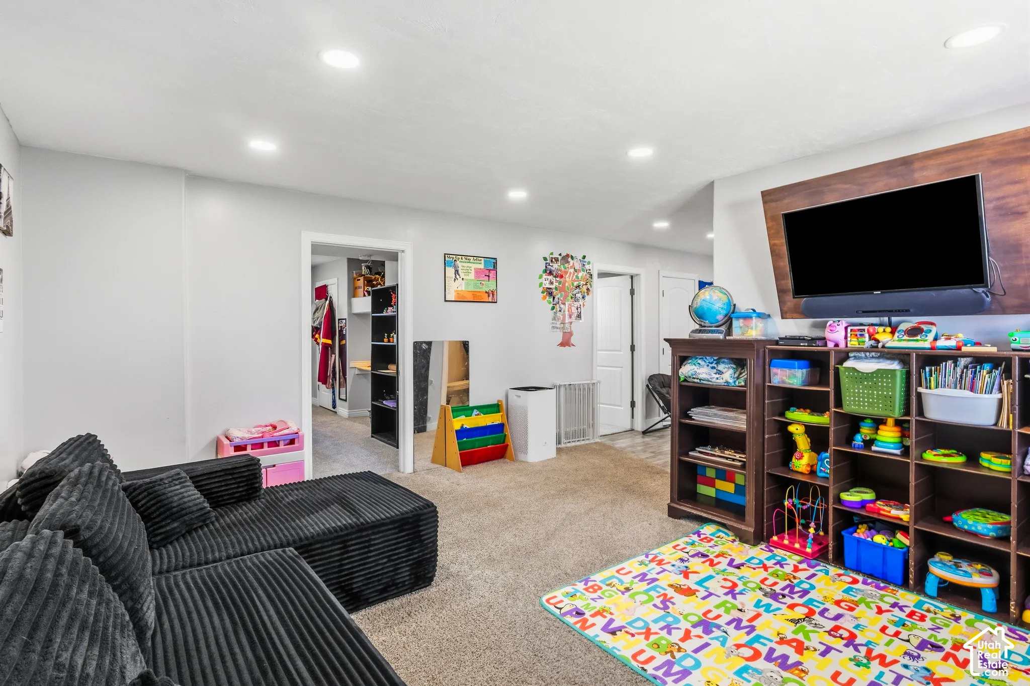 Recessed ceiling lights, neutral carpet flooring, wood-finish built-in shelving, and a wall-mounted television on a wood-finish panel