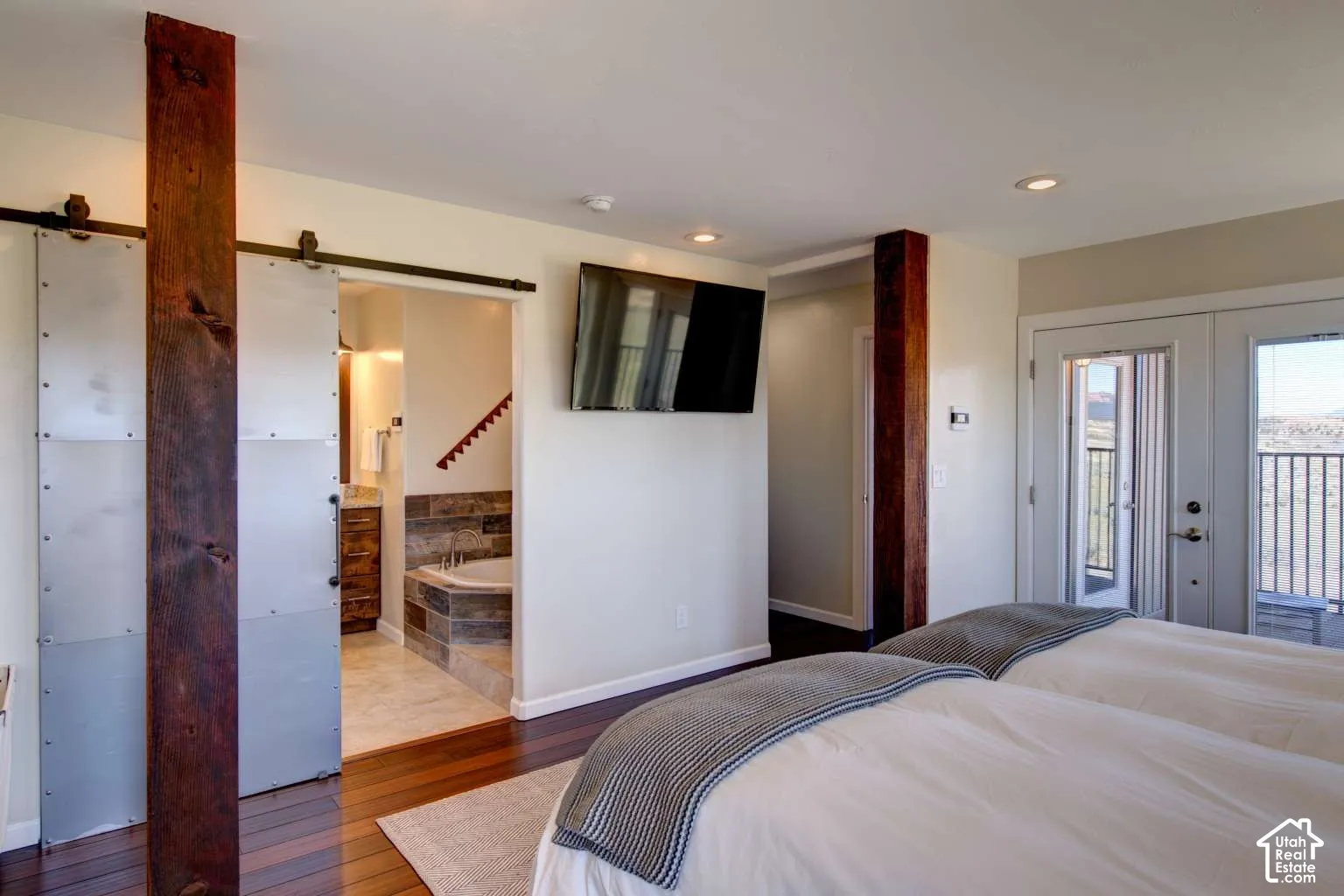 Hardwood flooring throughout, recessed ceiling lighting, a wall-mounted television, and a sliding barn door entry to the bathroom featuring a built-in tub with tile surround