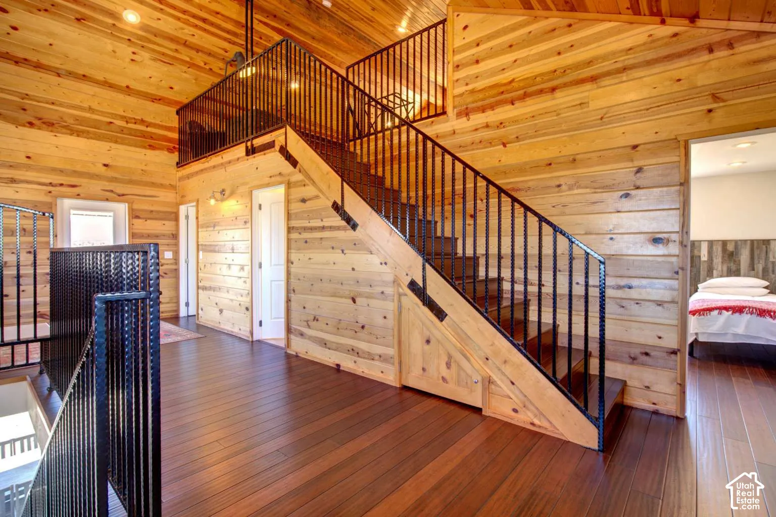 Wood-paneled walls and ceiling, dark wood-finish flooring, staircase with black metal railings, recessed lighting, and wall sconces