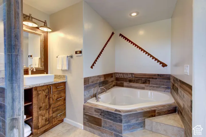 Built-in corner soaking tub with tile surround, wood-finish vanity with countertop, vessel sink, and overhead vanity lighting