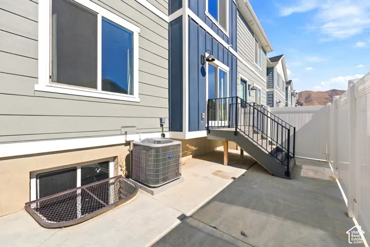 Gray horizontal siding with blue vertical accents, concrete patio, white privacy fence, exterior staircase with black railings, and a basement window well with a security grate