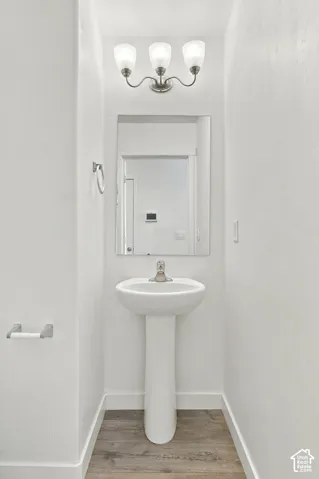 Pedestal sink, wall-mounted rectangular mirror, three-light vanity fixture, wood-finish flooring, and a towel ring