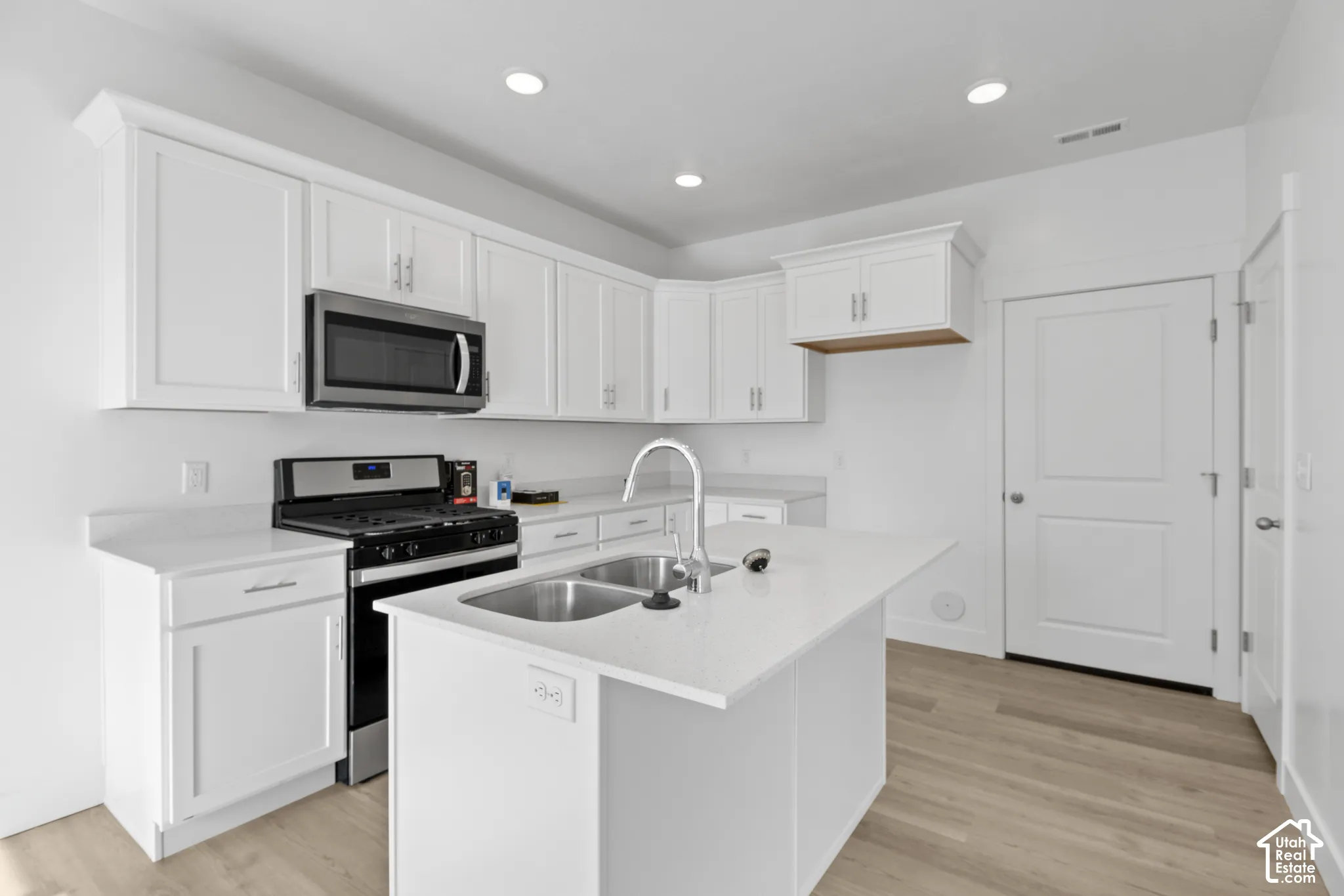 Kitchen featuring white shaker-style cabinetry, stainless steel appliances, a center island with an undermount dual-basin sink, light-toned wood-finish flooring, and recessed ceiling lighting