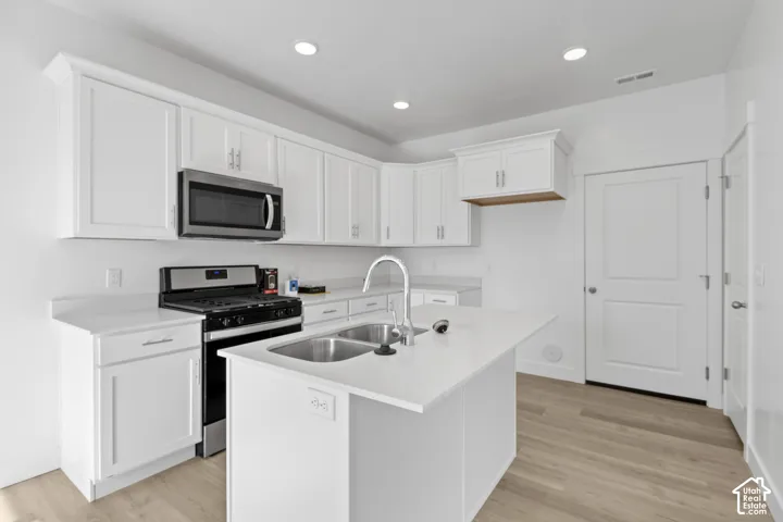 Kitchen featuring white shaker-style cabinetry, stainless steel appliances, a center island with an undermount dual-basin sink, light-toned wood-finish flooring, and recessed ceiling lighting