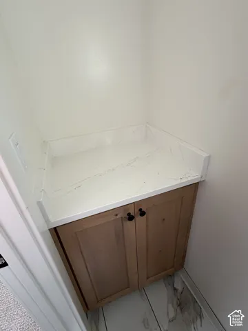 Wood-finish cabinetry with matte black hardware, white stone countertop, white walls, and stone-finish tile flooring