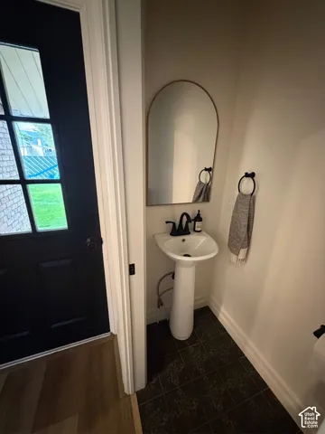Half bath next to back door. Pedestal sink with matte black faucet, arched mirror, dark tiled flooring, and wood-finish flooring