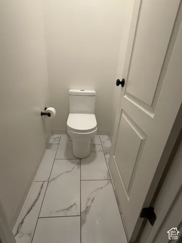 White porcelain toilet with black toilet paper holder, set against white walls and white tile flooring featuring grey veining