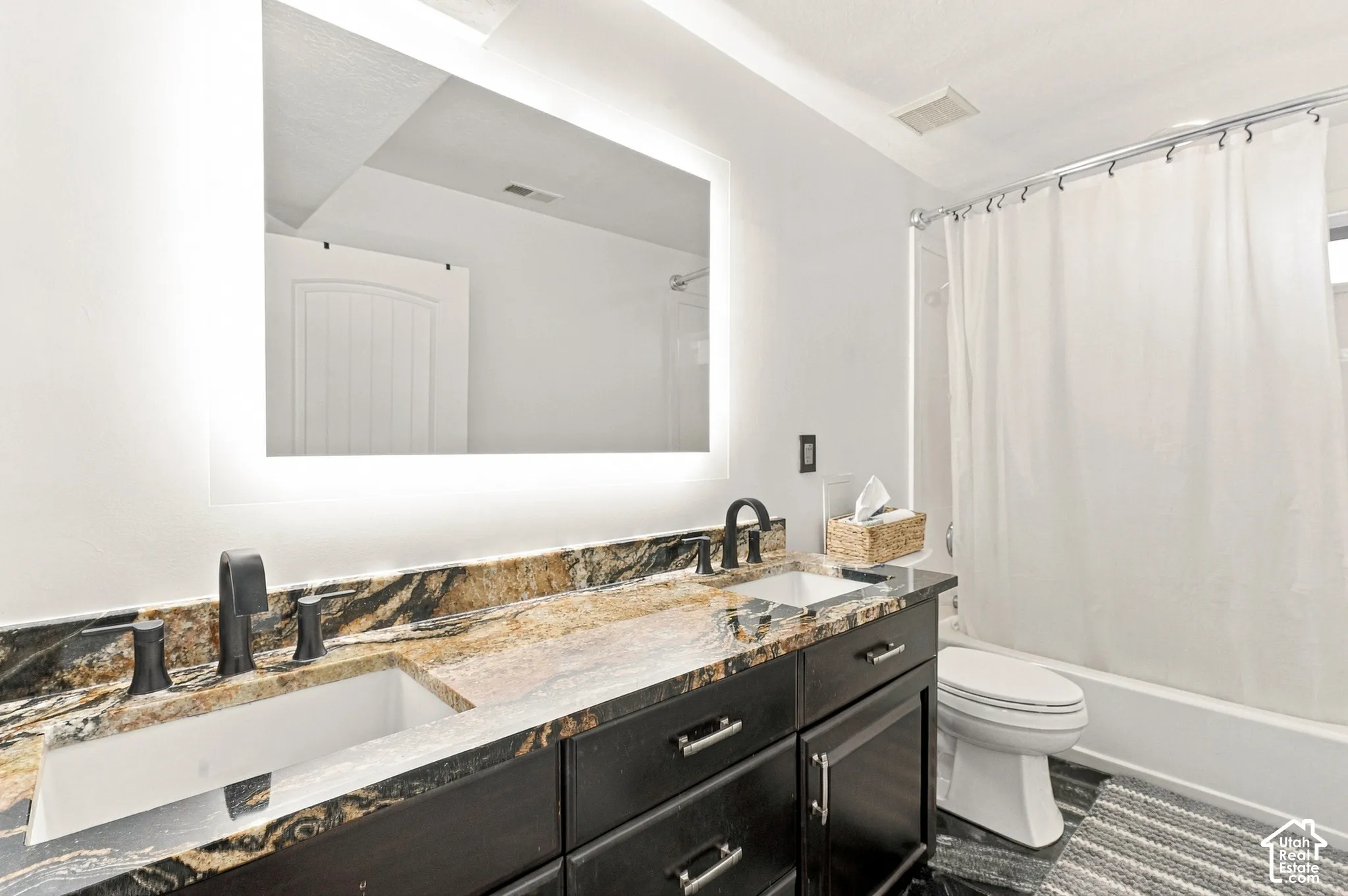 Double vanity with integrated sinks, back-lit mirror, dark wood-finish cabinetry with silver-tone hardware, and a stone countertop